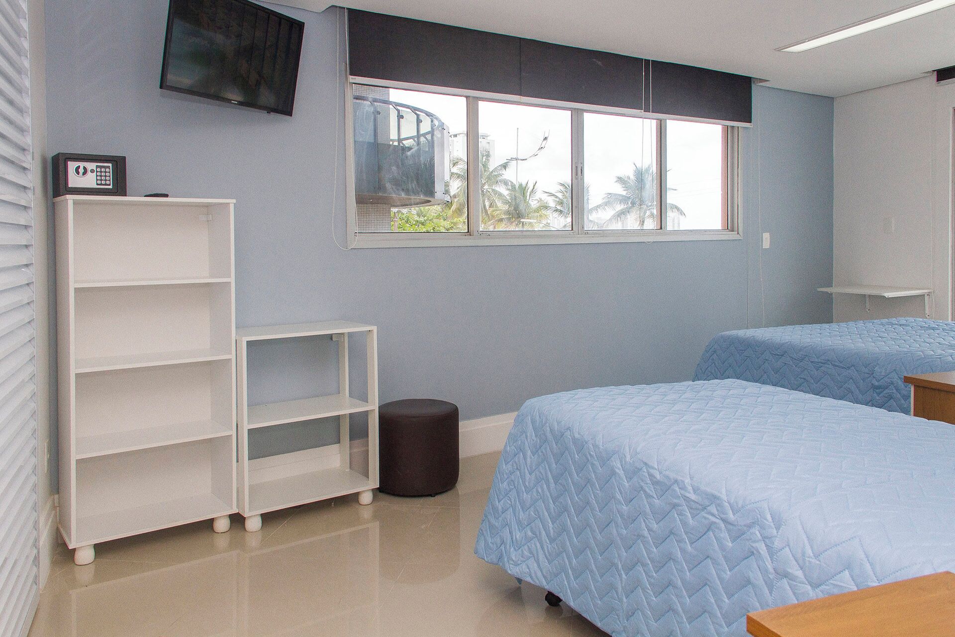 A bedroom with two beds, a white shelving unit, a small round stool, and a window with a view of palm trees.