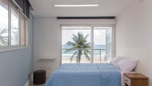 Une chambre à coucher comprenant un lit, une table de chevet, un petit tabouret et une grande fenêtre donnant sur la plage et les palmiers.