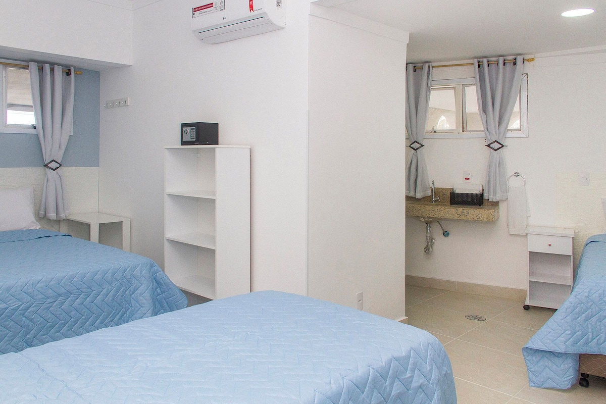 A hotel room with two beds, a small white shelf, a mirror above the sink, and a window with curtains.