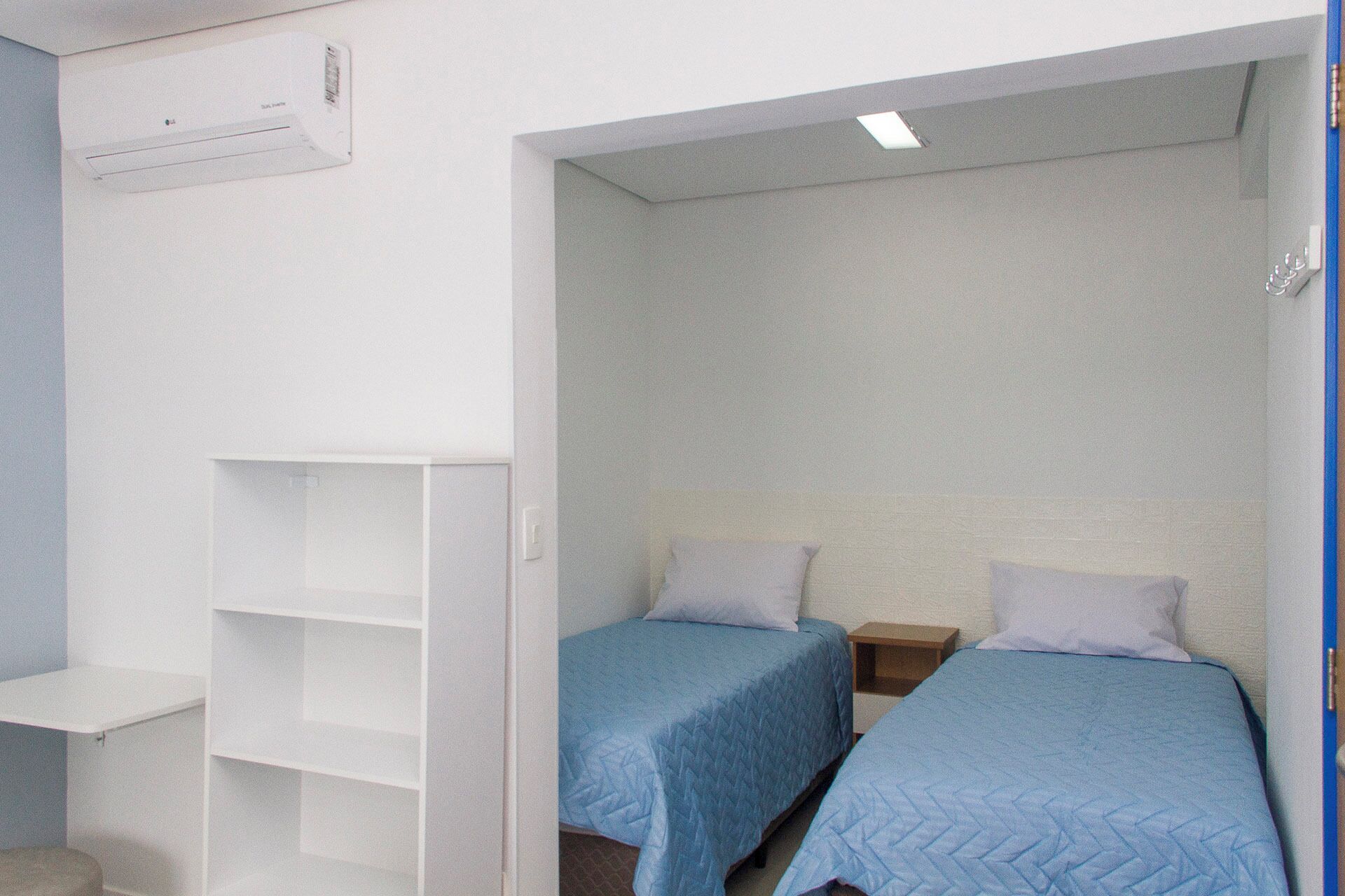 A small, compact room with two single beds, a white shelving unit, and a ceiling-mounted air conditioning unit.