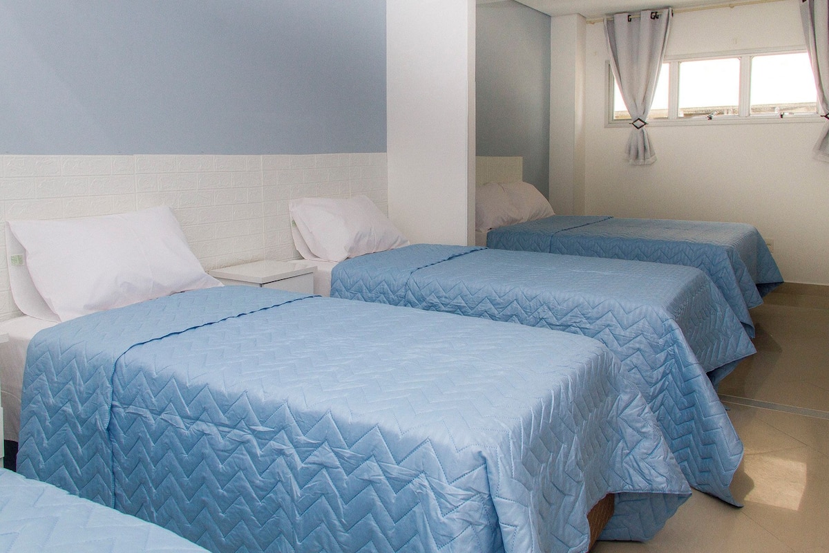 A hotel room with three beds, each with a blue quilted bedspread. There is a white headboard and a small bedside table with a lamp.