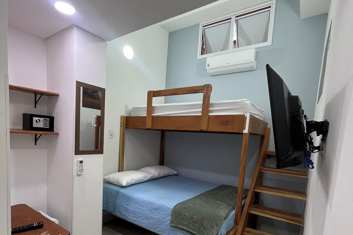 A compact room with a bunk bed, a desk with a chair, a small stool, and a TV mounted on the wall.