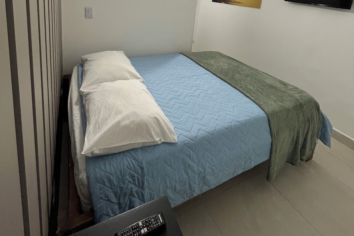 A single bed with a blue quilt, white pillows, and a green blanket. There is a black TV on the wall and a remote control on a small table.