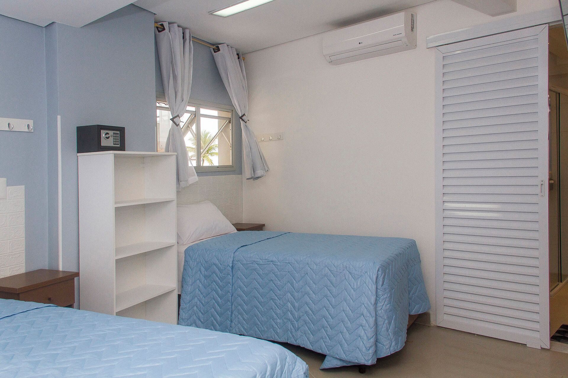 A bedroom with two beds, a wardrobe, a shelf unit, and a window with curtains.