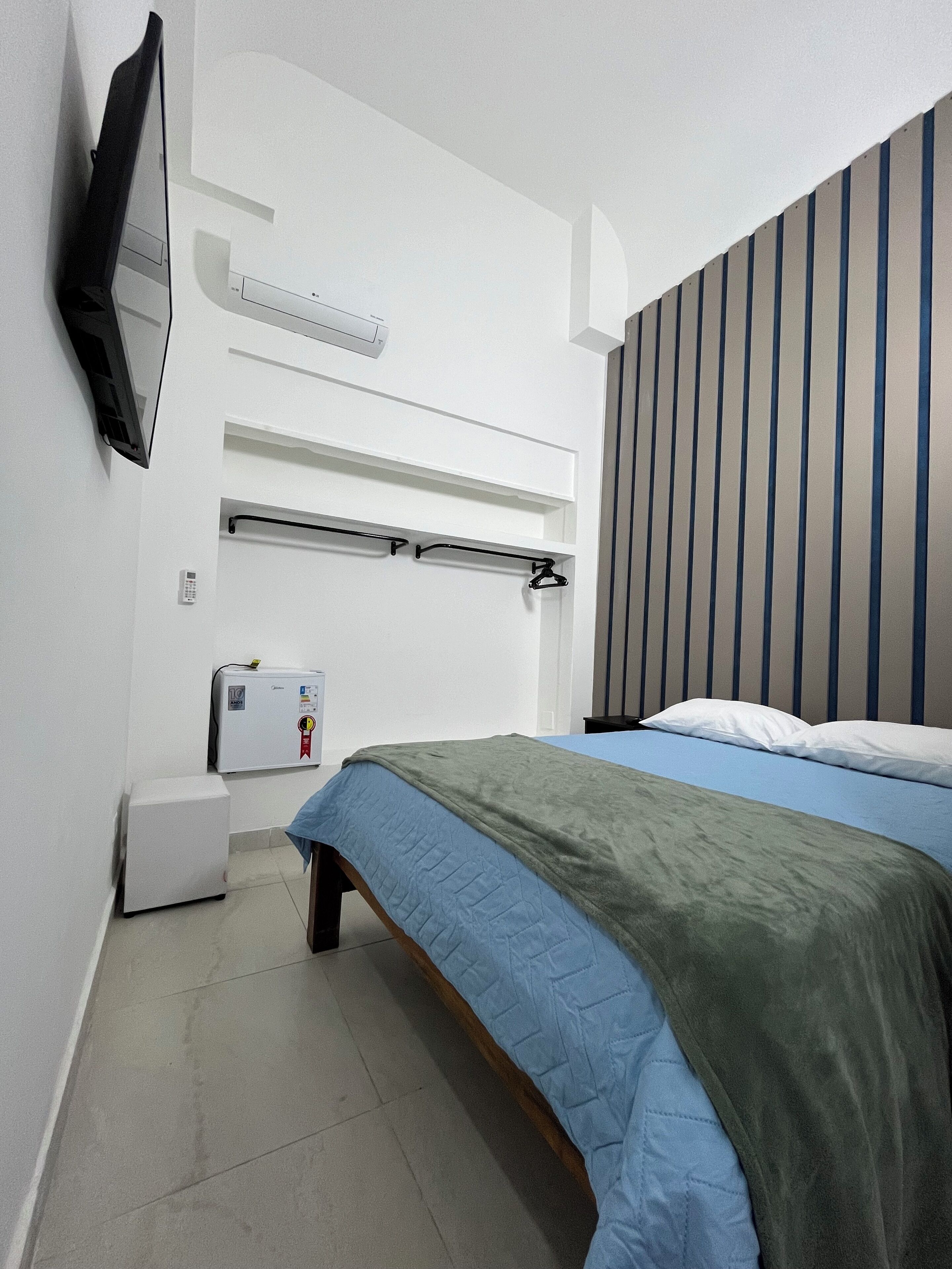 A bedroom with a bed, a television mounted on the wall, a small refrigerator, and a window with vertical blinds.