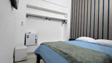 Un dormitorio con cama, televisor montado en la pared, refrigerador pequeño y ventana con persianas verticales.
