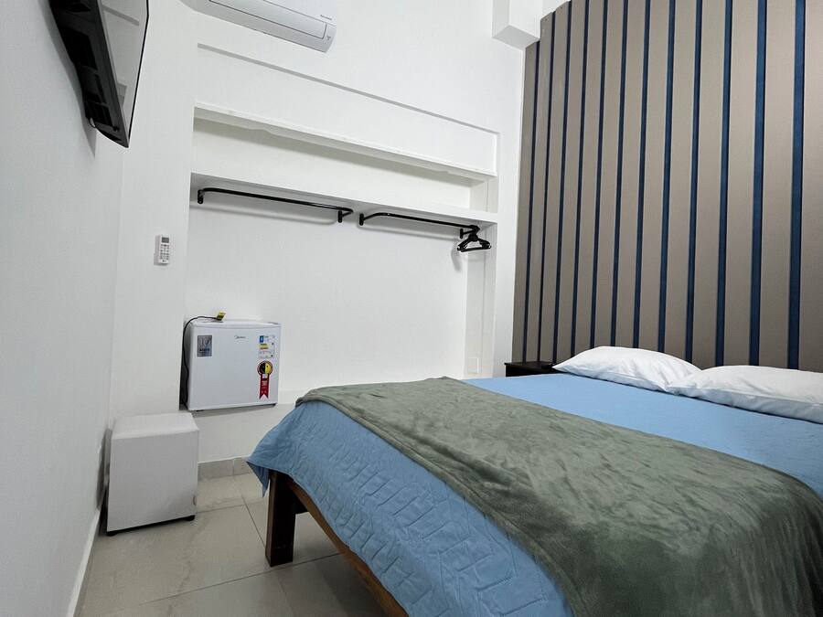 Un dormitorio con cama, televisor montado en la pared, refrigerador pequeño y ventana con persianas verticales.