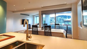 A modern hotel room with a large window offering a city view, a dining area with a table and chairs, and a kitchen area with a sink and faucet. - U Suites on Oriental Parade (Wellington)