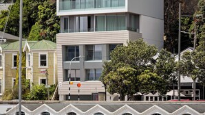 A modern building with large windows and a flat roof, situated near a body of water with a sailboat docked nearby. - U Suites on Oriental Parade (Wellington)