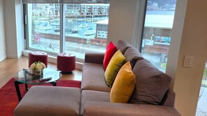 A modern living room with a large window, a flat-screen TV, a glass coffee table, and a sofa with colorful cushions. - U Suites on Oriental Parade (Wellington)
