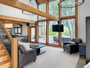 Amplia sala de estar con escalera, televisor grande y vistas al exterior.