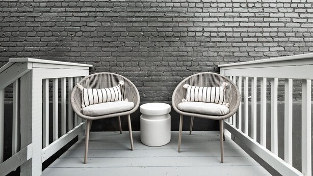 Un balcon avec deux fauteuils en osier et une petite table ronde, le tout adossé à un mur de briques.