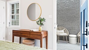 Une chambre à coucher avec une commode en bois, un miroir rond, une couvre-lit verte et une fenêtre avec des volets blancs.