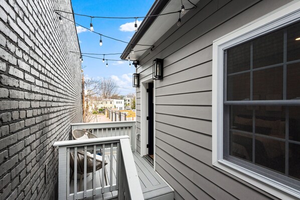 A narrow outdoor corridor with a brick wall on one side and a gray siding house on the other. There is a small porch with a railing and a few chairs.