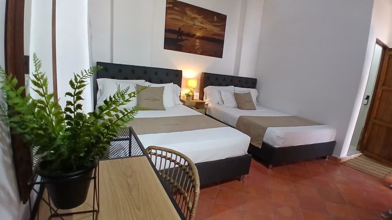 A hotel room with two beds, a wooden table, a potted plant, and a painting on the wall.