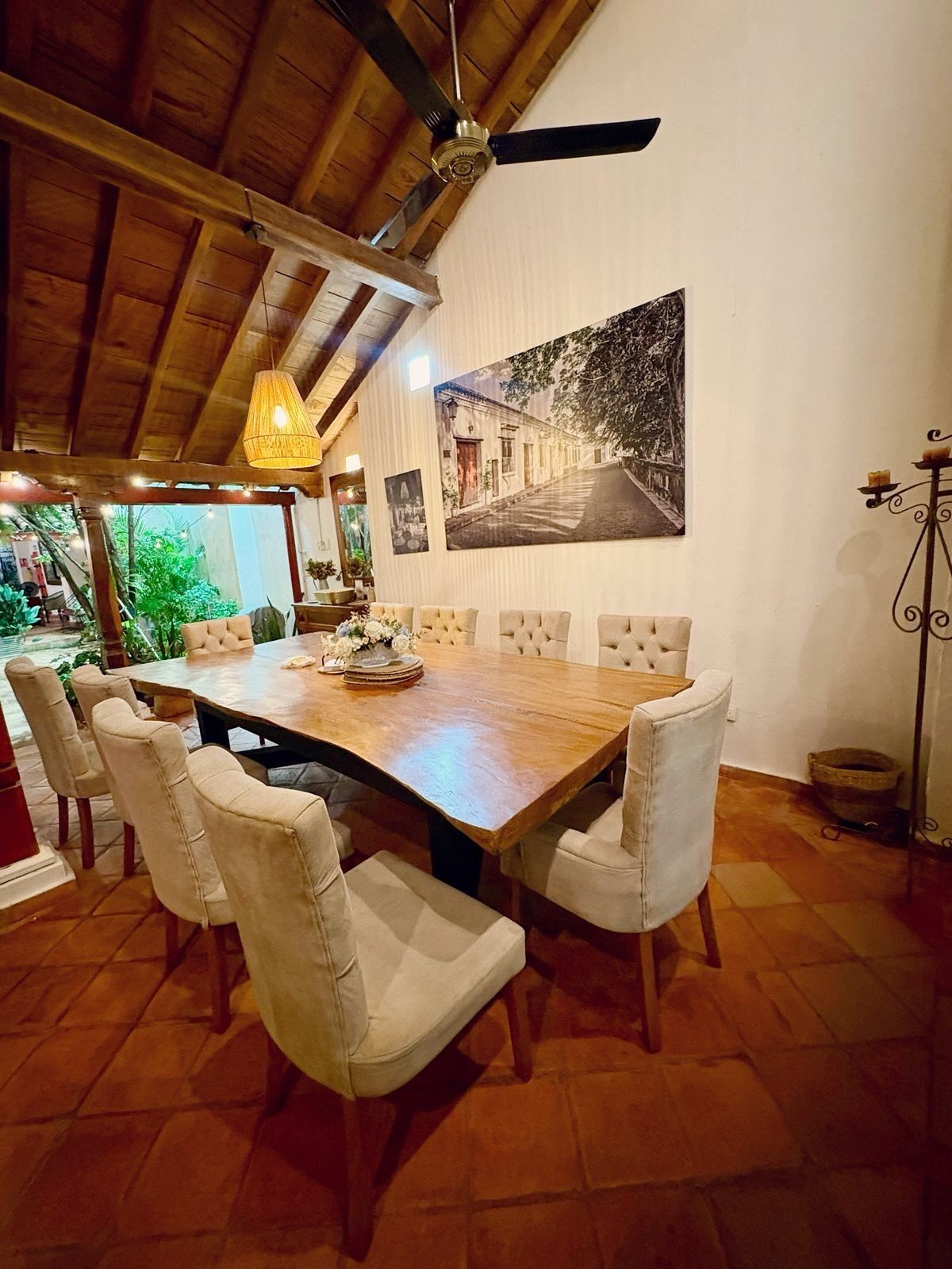 Un comedor con mesa de madera, sillas acolchadas, ventilador de techo y un cuadro en la pared.