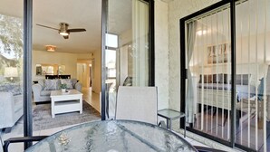 Un salon moderne avec une table en verre, un ventilateur de plafond et une vue sur une chambre à coucher, accessible par une porte-fenêtre coulissante.