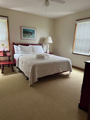 A neatly made bed with a white comforter and pillows, a bedside table with a lamp, a framed picture on the wall, and a window with blinds.
