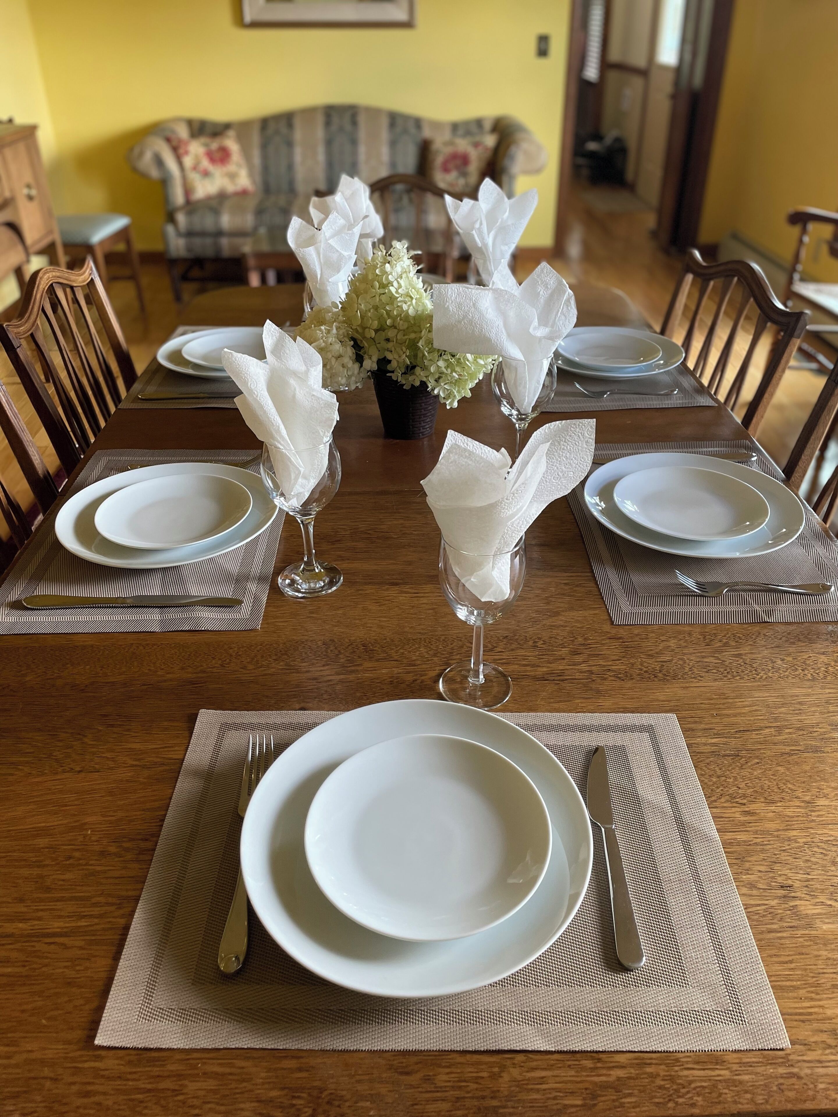 Una mesa de comedor dispuesta con platos blancos, vasos y cubiertos, adornada con un arreglo floral en el centro.