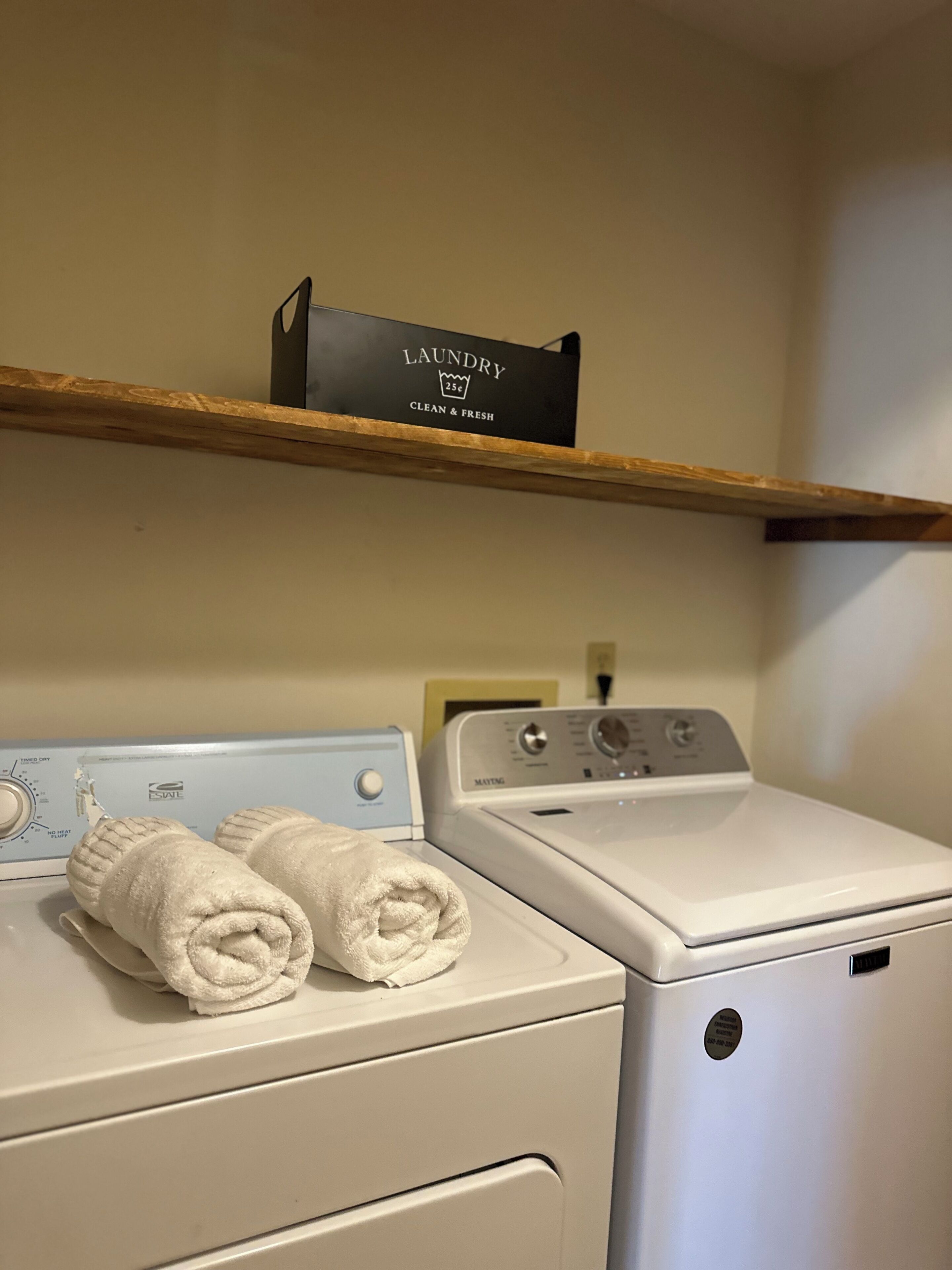 A laundry area with a washing machine and dryer, a shelf with a laundry bag, and a wall-mounted thermostat.