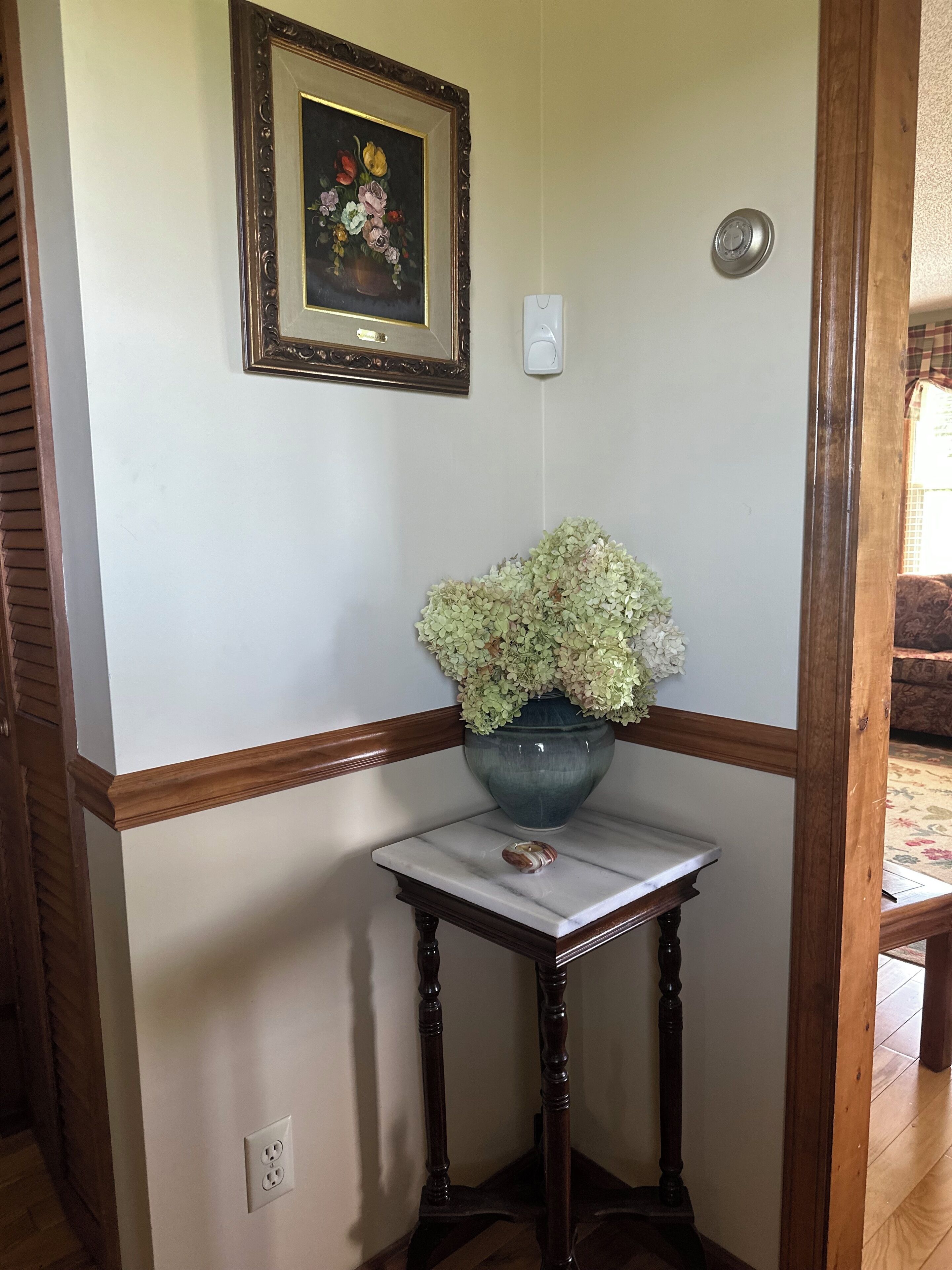 Canto de um quarto com um banco de madeira, uma pequena mesa com um vaso de flores, um quadro na parede e um interruptor de luz.