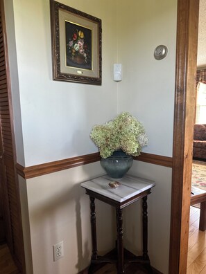 A corner of a room with a wooden bench, a small table with a vase of flowers, a framed picture on the wall, and a light switch.