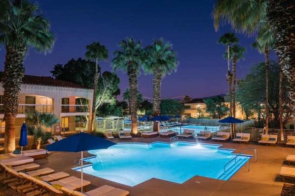 A swimming pool with lounge chairs and umbrellas, surrounded by palm trees and a building with balconies. - Ramada Palm Springs (Palm Springs)