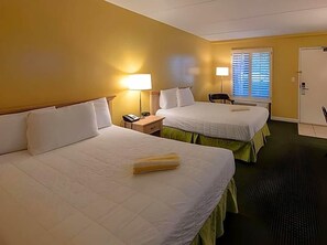Ein Hotelzimmer mit zwei Betten, einem Schreibtisch, einem Stuhl, einer Lampe und einem Fenster mit Jalousien.