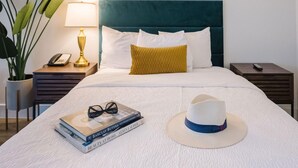 Una cama bien hecha con una colcha blanca, un cabecero azul verdoso y una sola almohada color mostaza. Hay una pila de libros y un sombrero blanco sobre la cama.