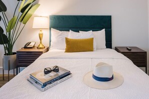 A neatly made bed with a white comforter, a teal headboard, and a single mustard-colored pillow. A stack of books and a white hat are placed on the bed. - Ramada Palm Springs (Palm Springs)