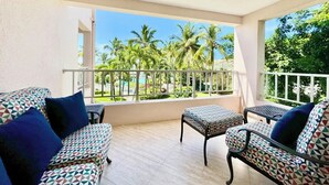 A balcony with a patterned sofa and ottoman, a small table, and a view of palm trees and greenery.