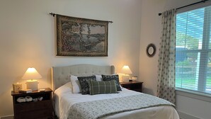A neatly made bed with a white comforter and multiple pillows, flanked by two bedside tables with lamps and a small nightstand with a tissue box and book. A decorative tapestry hangs above the bed, and a window with blinds is visible to the right.
