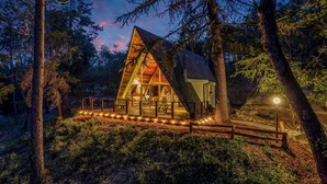 Una cabaña de techo triangular, rodeada de árboles, e iluminada por luces de cadena.