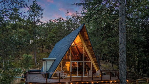 Una cabaña de madera triangular con frente de vidrio, rodeada de árboles y una terraza con luces.
