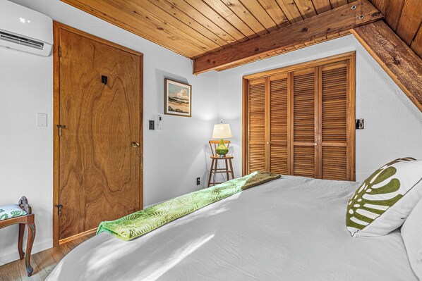 A bedroom with a wooden ceiling, a bed with a green and white patterned bedspread, a wooden wardrobe, and a small table with a lamp.