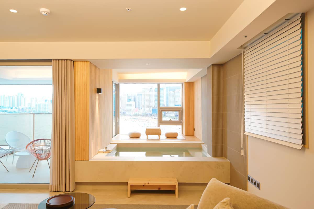 Una sala de estar moderna con vistas a un paisaje urbano a través de amplios ventanales, un rincón de descanso con sofá y sillones, y una pequeña mesa de madera.