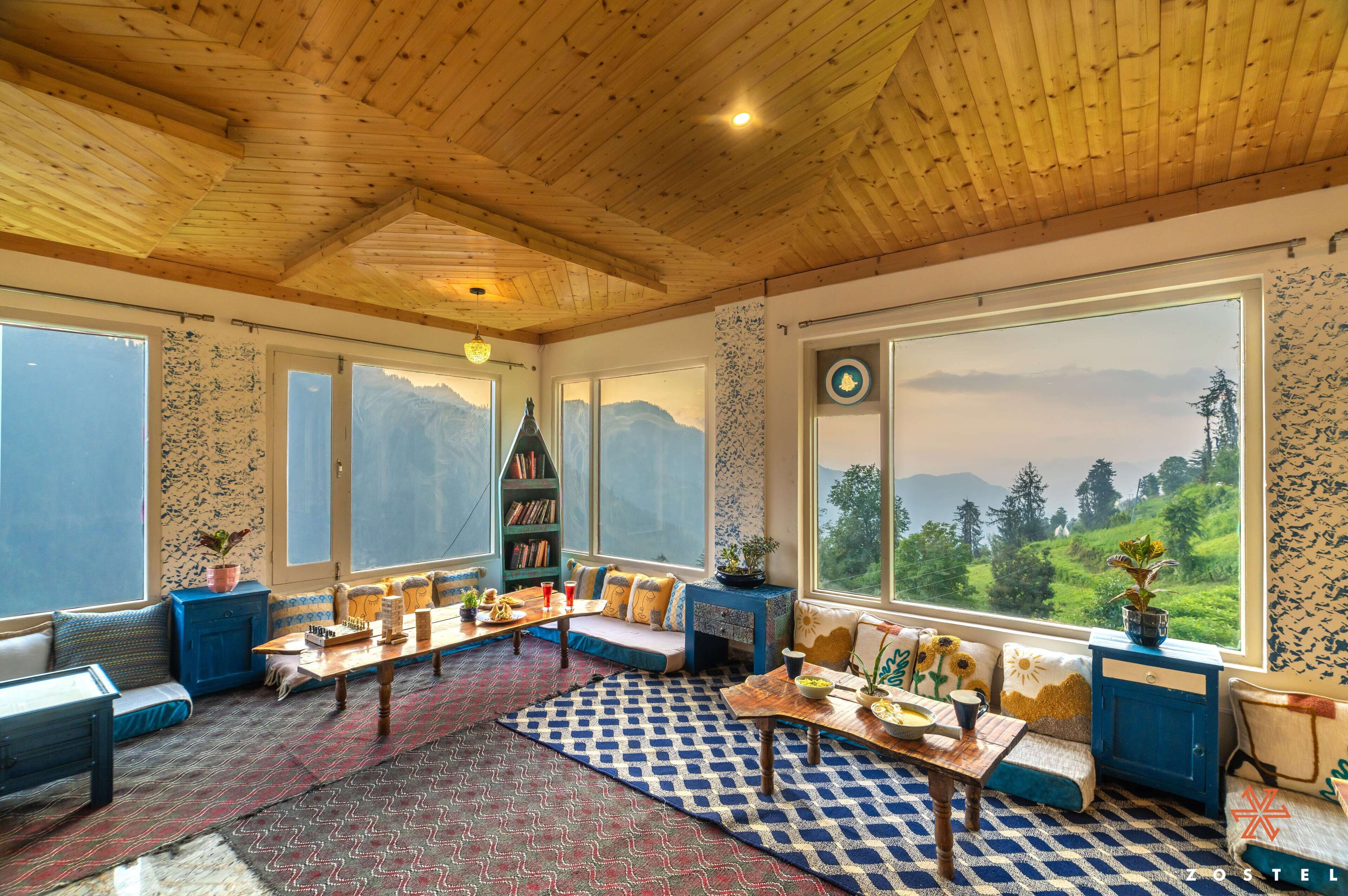 A cozy room with a wooden ceiling, large windows with a mountain view, a sofa, a table, and a bookshelf.
