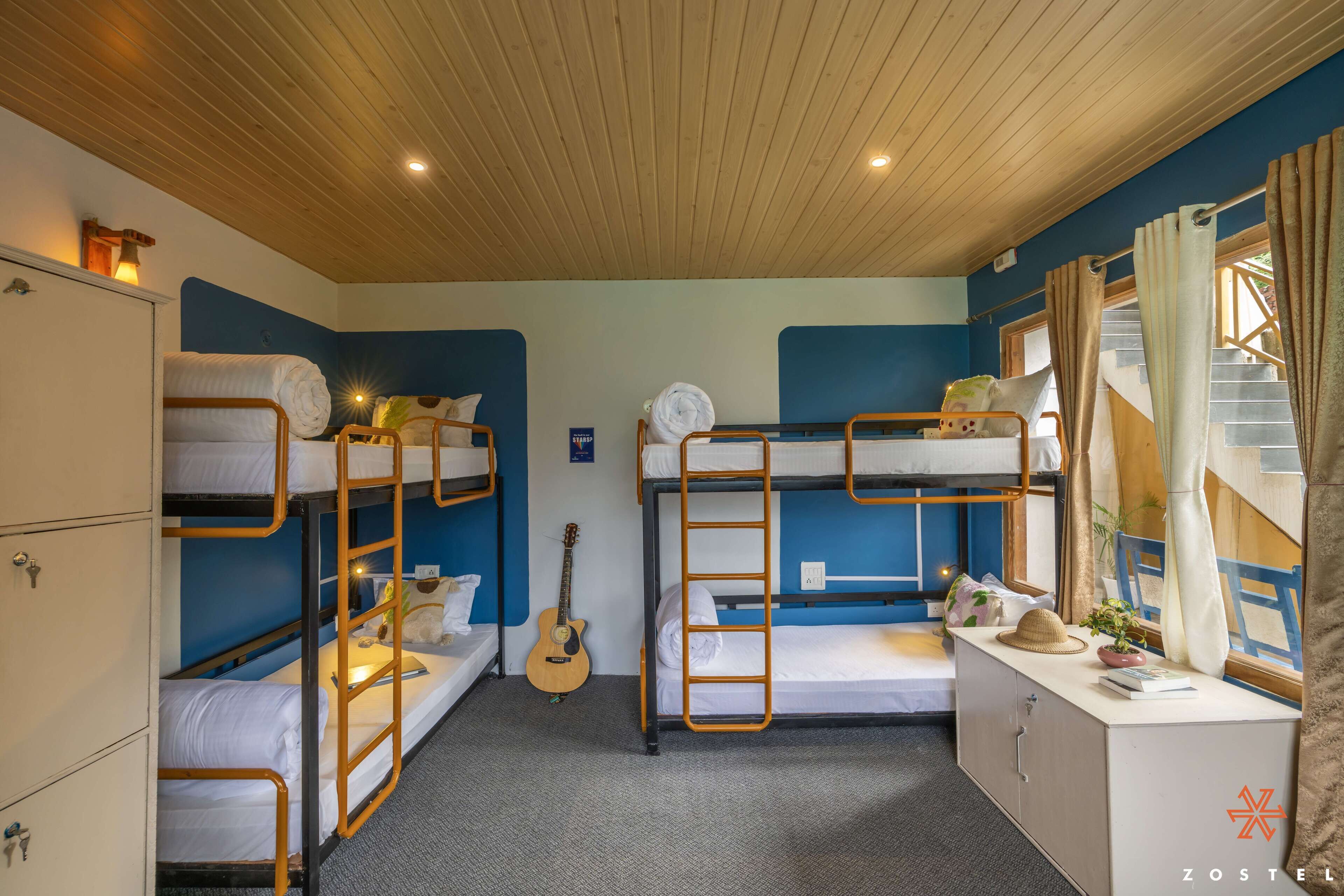A dormitory room with bunk beds, a guitar, a desk with a plant, and a staircase visible through the window.