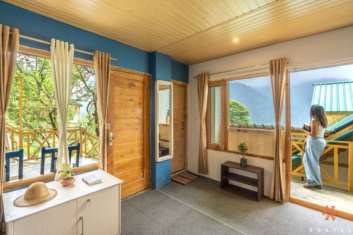 Ein Zimmer mit Holzbalkendecke, einem Schreibtisch mit Hut und Pflanze, einer Bank und Blick auf eine Holzterrasse sowie Berge durch große Fenster.