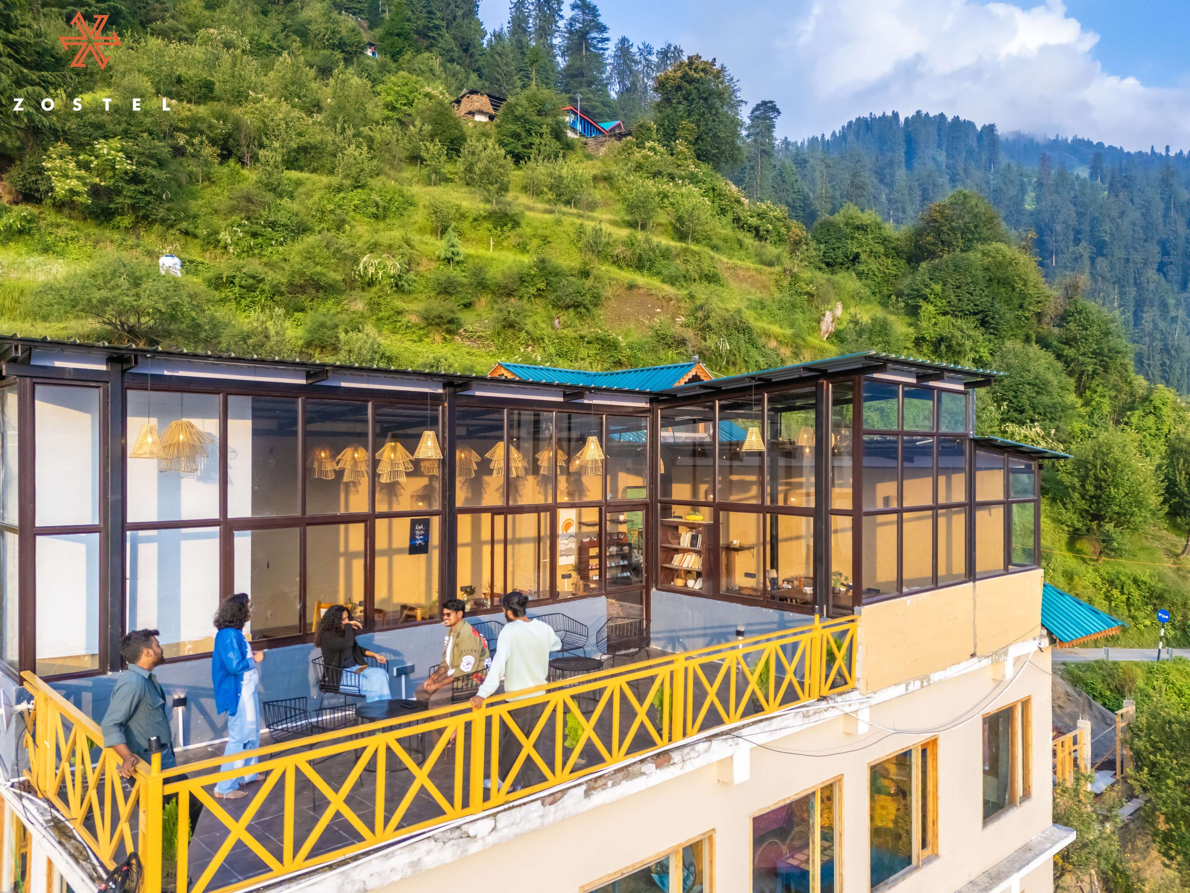 A modern building with large glass windows and a yellow railing, overlooking a lush green hillside.