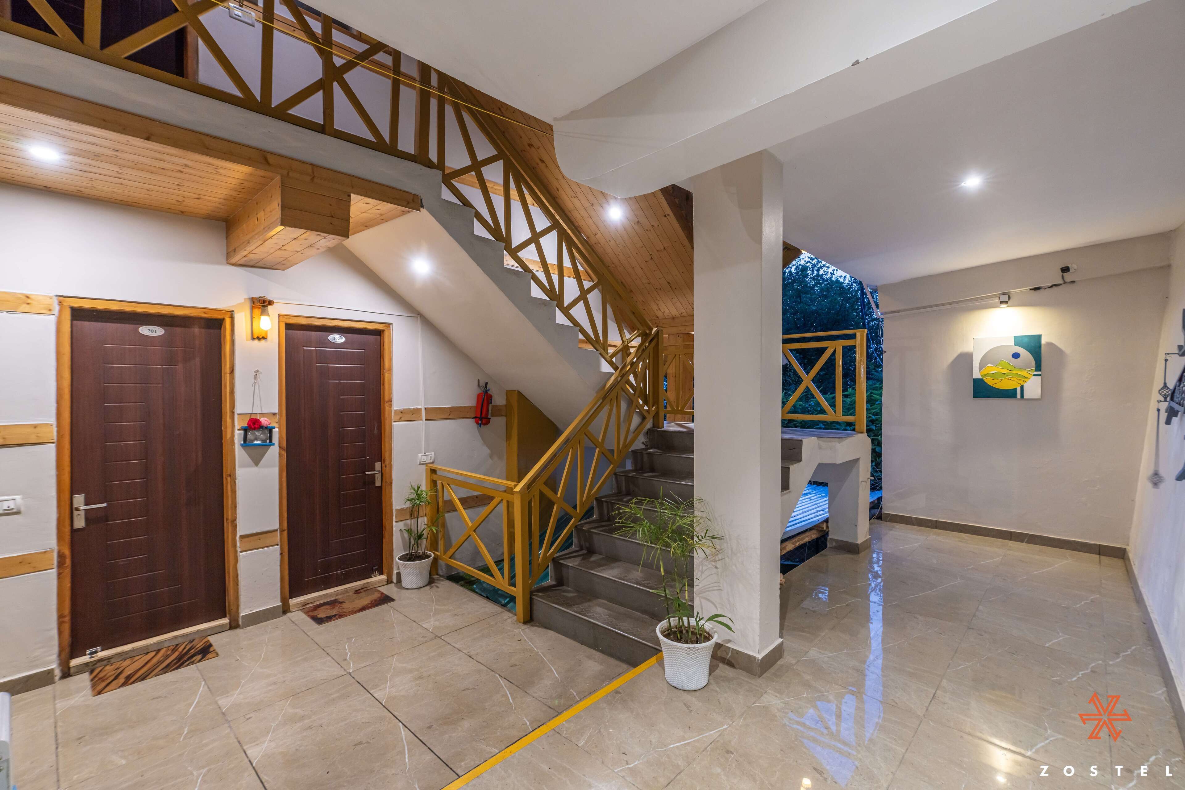Un hall d’hôtel avec un escalier en bois, deux portes, des plantes en pot et un lit en arrière-plan.