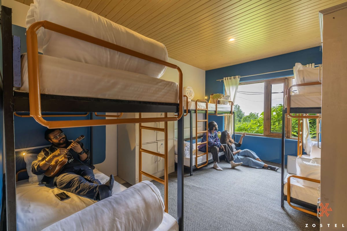 Ein Schlafsaalzimmer mit Etagenbetten, einem Fenster mit Blick auf Grünflächen und einer Person, die Gitarre spielt.