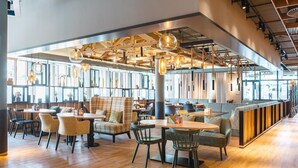 A modern restaurant with wooden flooring, multiple seating areas including tables and booths, hanging pendant lights, and large windows allowing natural light. - aja Garmisch-partenkirchen (Garmisch-Partenkirchen)