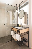 A modern bathroom with a glass shower enclosure, a wooden vanity with a black countertop, a round mirror, and a window providing natural light.