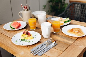 Una mesa de desayuno con platos de comida, una taza, vasos de zumo de naranja y una jarra de leche.