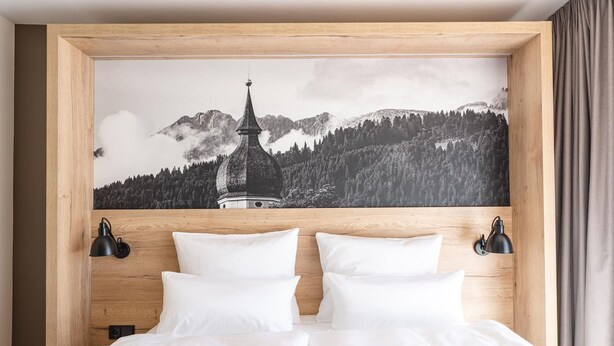 Une chambre à coucher avec une tête de lit en bois, un lit recouvert de draps blancs, deux lampes murales noires et un tableau représentant un paysage de montagne au-dessus du lit.