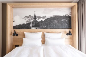 Une chambre à coucher avec une tête de lit en bois, un lit recouvert de draps blancs, deux lampes murales noires et un tableau représentant un paysage de montagne au-dessus du lit.