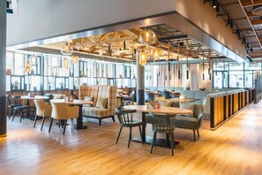 A modern restaurant with wooden flooring, multiple seating areas including tables and booths, hanging pendant lights, and large windows allowing natural light. - aja Garmisch-partenkirchen (Garmisch-Partenkirchen)