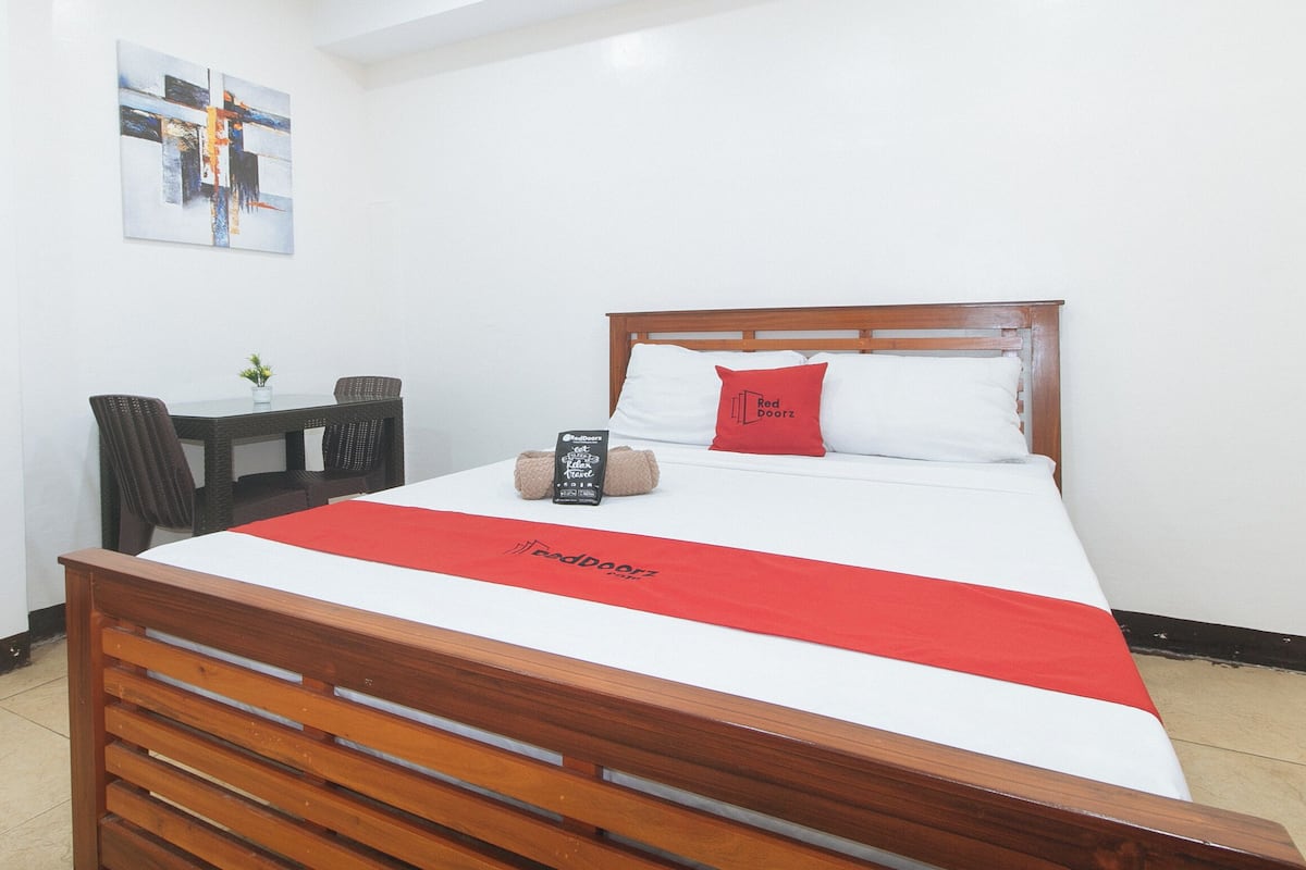 Habitación de hotel con cama de madera, ropa de cama blanca, una manta roja, almohada Red Doorz, una mesita con planta y un cuadro en la pared.