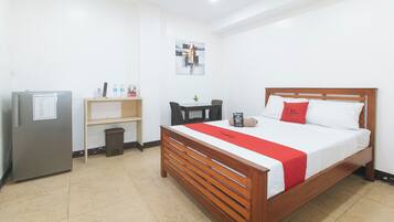 Quarto de hotel com uma cama, cabeceira de madeira, almofada vermelha, pequena mesa com uma planta, prateleira com objetos e um refrigerador.